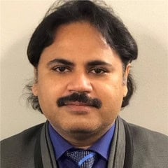 A man with dark hair, a mustache, and a light beard is wearing a suit jacket, blue shirt, and patterned tie, posing against a plain background.