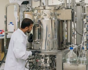 A Mines doctoral student in a lab coat and gloves inspects a large stainless steel bioreactor, surrounded by bottles and lab equipment. The setup, hinting at sustainable agricultural bioplastics research, features various knobs and gauges in the bustling laboratory environment. - BioWRAP