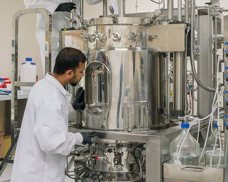 A Mines doctoral student in a lab coat and gloves inspects a large stainless steel bioreactor, surrounded by bottles and lab equipment. The setup, hinting at sustainable agricultural bioplastics research, features various knobs and gauges in the bustling laboratory environment. - BioWRAP