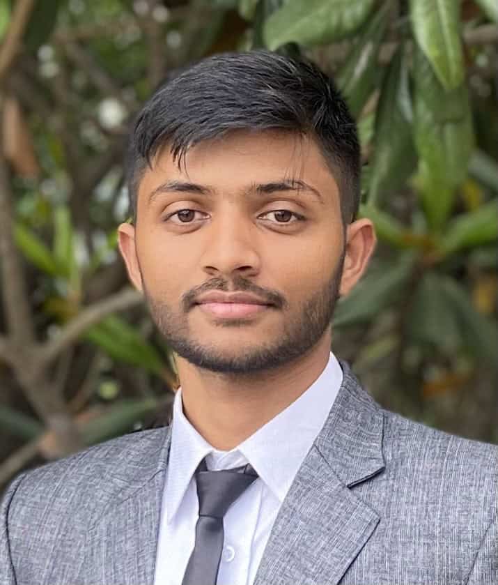 A person with short dark hair and a trimmed beard is wearing a gray suit, white shirt, and black tie. They are standing outdoors with green foliage in the background, looking directly at the camera with a slight smile.