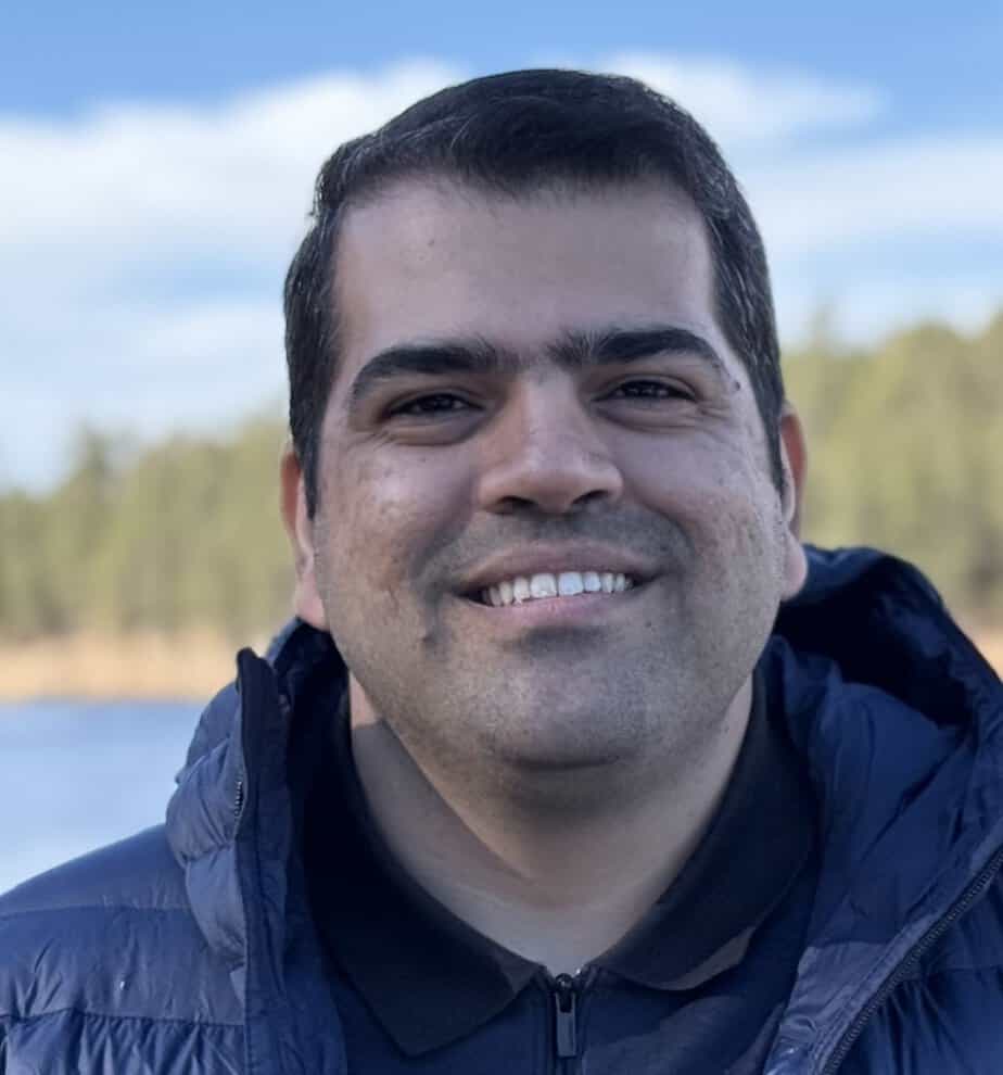 A person with short dark hair. They are wearing a dark jacket and a black shirt. The background features a blurred landscape with trees and a body of water under a blue sky.