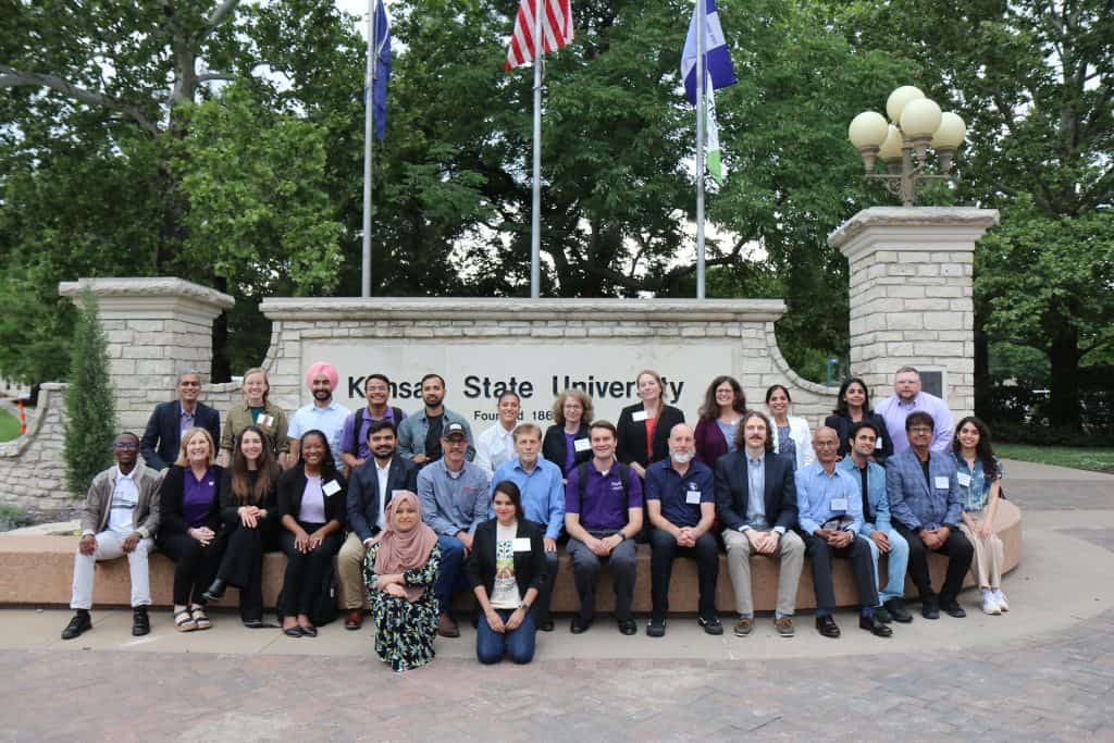 A diverse group of people poses together in front of a stone Kansas State University sign and flagpoles during the 2023 BioWRAP annual meeting, with trees in the background. Some are sitting on a bench, others standing behind them, all smiling at the camera. - BioWRAP
