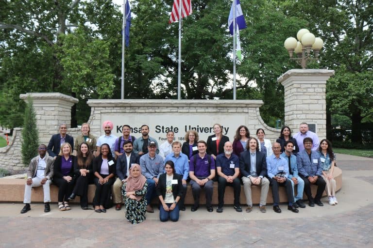 A diverse group of people poses together in front of a stone Kansas State University sign and flagpoles during the 2023 BioWRAP annual meeting, with trees in the background. Some are sitting on a bench, others standing behind them, all smiling at the camera. - BioWRAP