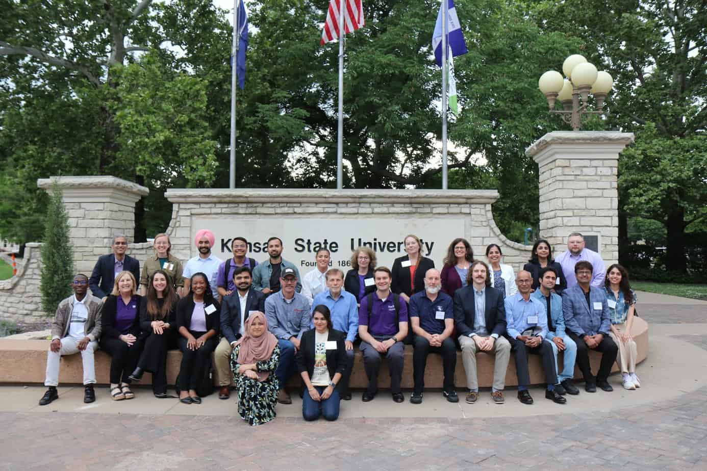 A diverse group of people poses together in front of a stone Kansas State University sign and flagpoles during the 2023 BioWRAP annual meeting, with trees in the background. Some are sitting on a bench, others standing behind them, all smiling at the camera. - BioWRAP