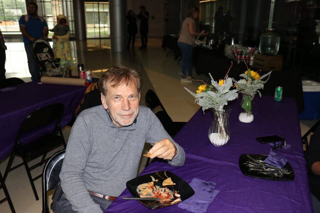 At the 2023 Annual Meeting, a man with gray hair sits at a table with a plate of food, holding a chip. The purple tablecloth and vases of artificial sunflowers brighten the scene, with other attendees and tables visible in the background. - BioWRAP