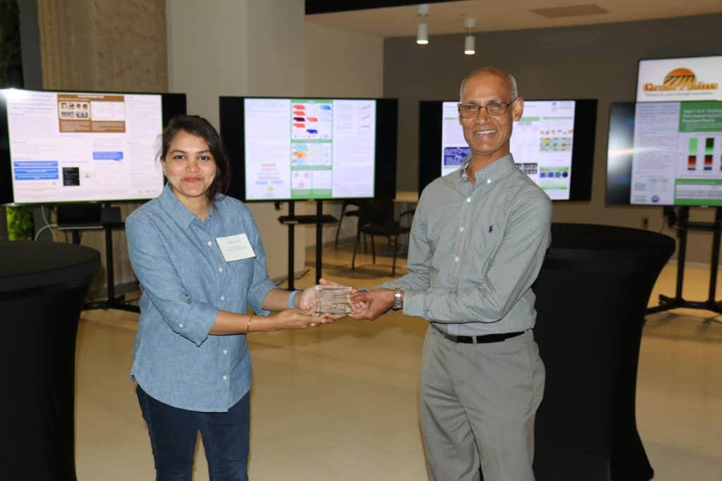 Two people stand indoors, smiling and holding a glass award together at the 2023 Annual Meeting. Behind them, multiple digital screens display colorful charts and BioWRAP posters. Both are dressed in casual business attire. - BioWRAP