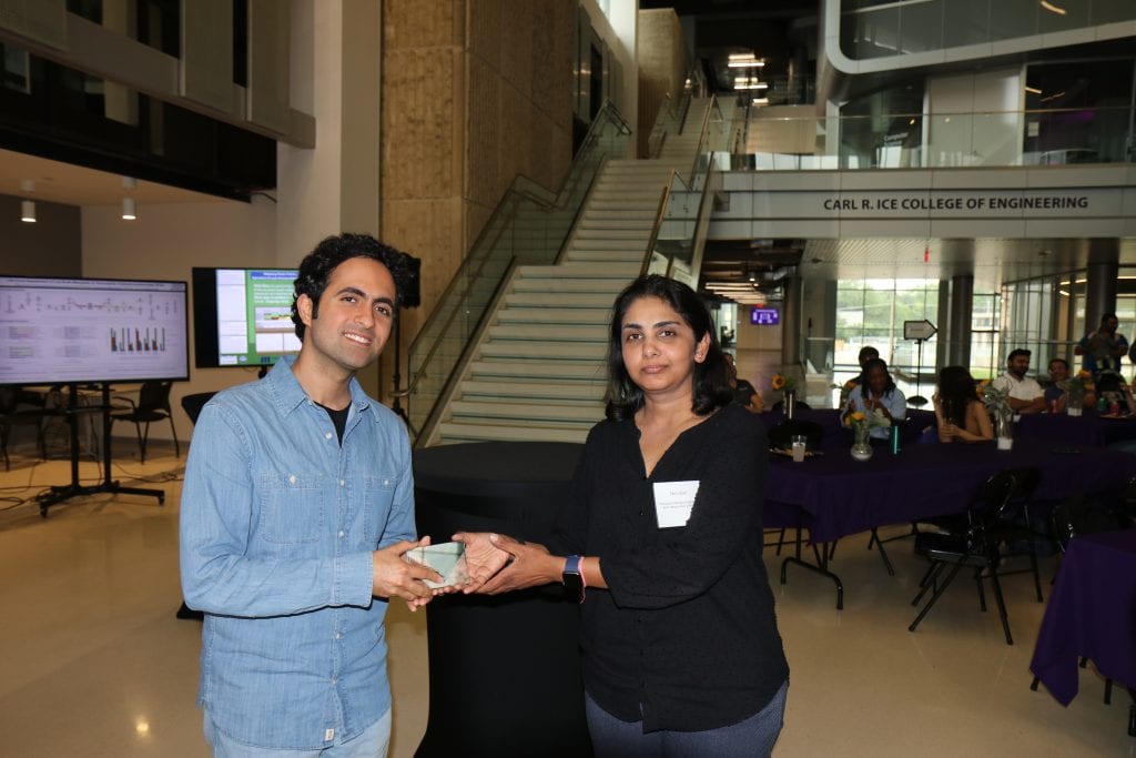 At the 2023 Annual Meeting, two people stand indoors, one handing an award to the other with smiles. Behind them is a staircase, a sign reading "Carl R. Ice College of Engineering," and people seated at tables for BioWRAP. - BioWRAP