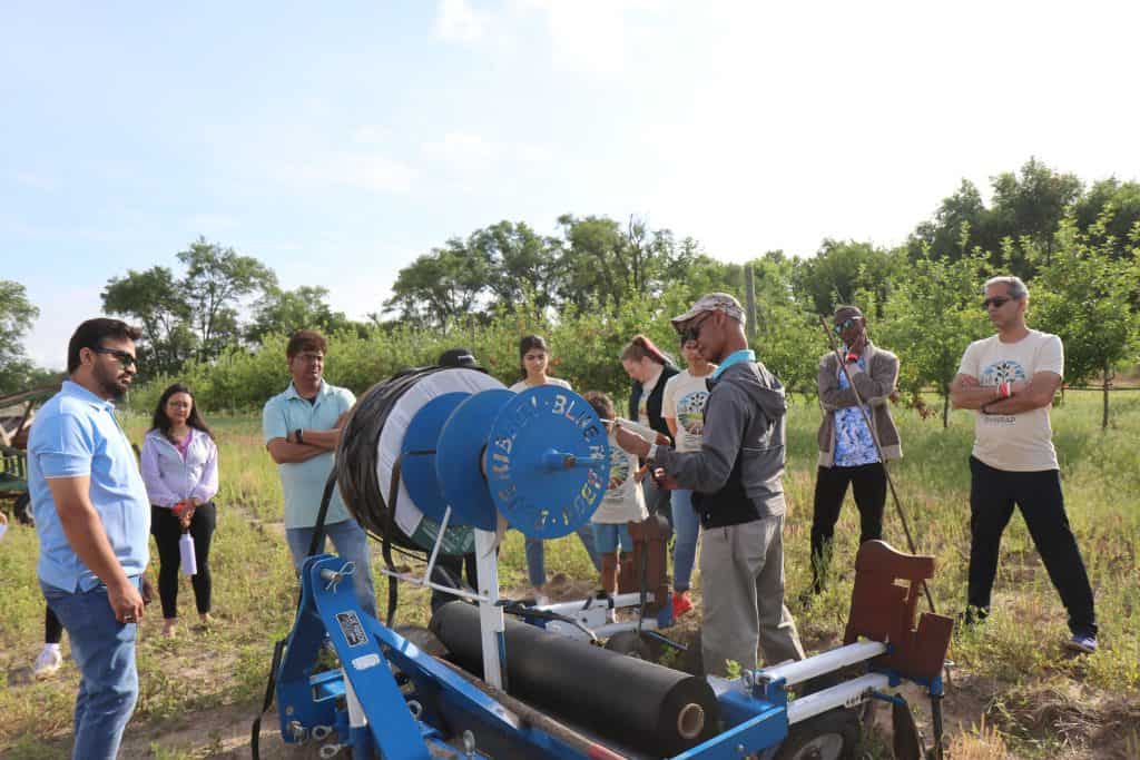 At the 2023 Annual Meeting, a group gathers outdoors around a blue and black BioWRAP agricultural machine, attentively listening as its features are explained in a grassy area with trees under a bright sky. - BioWRAP
