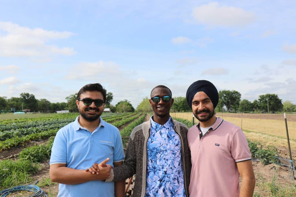 Three men stand together smiling in front of rows of crops at the 2023 BioWRAP Annual Meeting, enjoying the outdoor setting under a blue sky with scattered clouds. Casually dressed, they appear happy and relaxed on the farm field. - BioWRAP