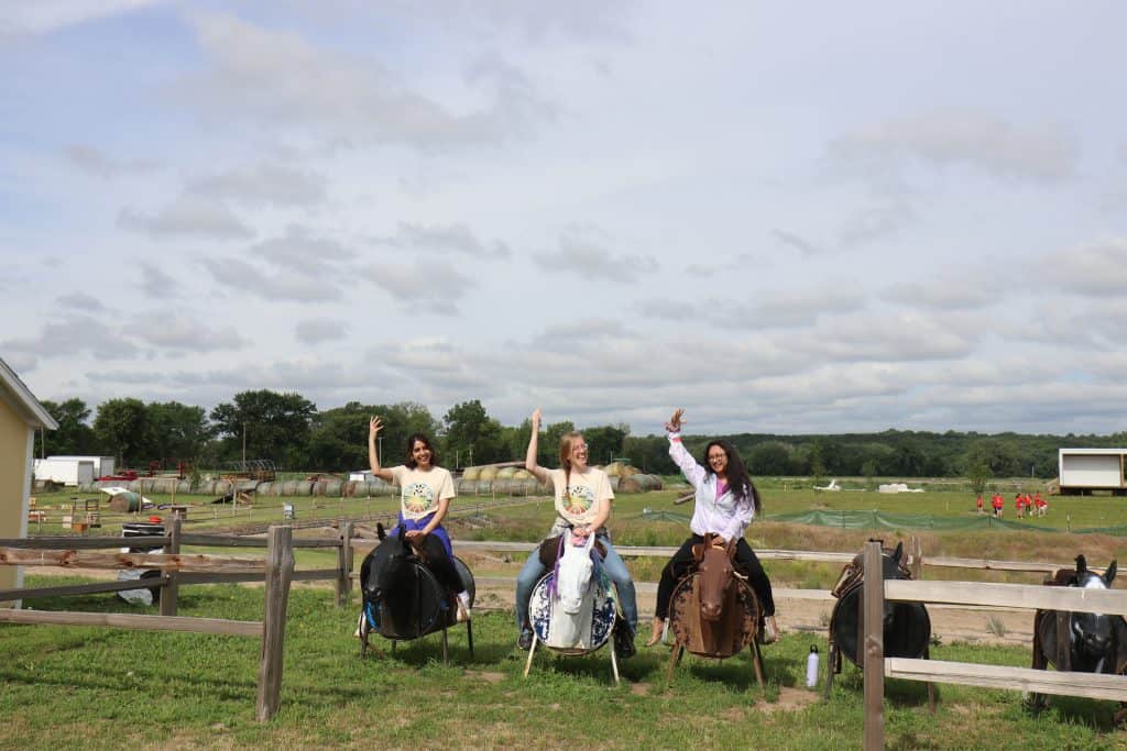 Three people sit on stationary saddles behind a wooden fence, waving and smiling at the camera during the 2023 BioWRAP Annual Meeting. The background shows a grassy field, scattered equipment, and a cloudy sky. - BioWRAP