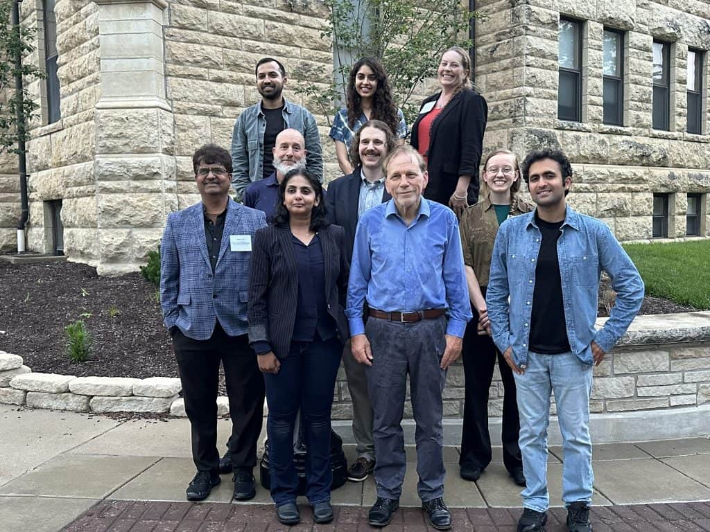 A group of ten people, diverse in age and gender, stand outside in front of a stone building at the 2023 BioWRAP Annual Meeting, smiling at the camera. Most are dressed in business-casual attire, with greenery and a tree behind them. - BioWRAP