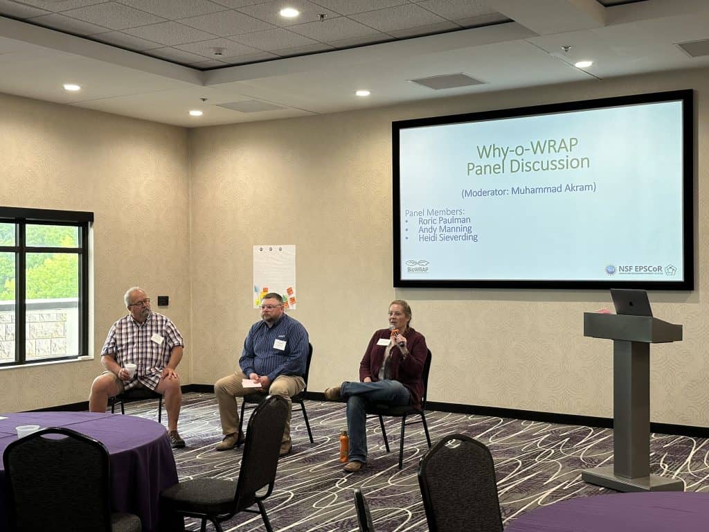 Three panelists sit at the front of a conference room for the "Why-o-WRAP Panel Discussion" at the 2023 BioWRAP Annual Meeting. A woman speaks into a microphone while two men listen. An empty podium and round tables are in the foreground. - BioWRAP