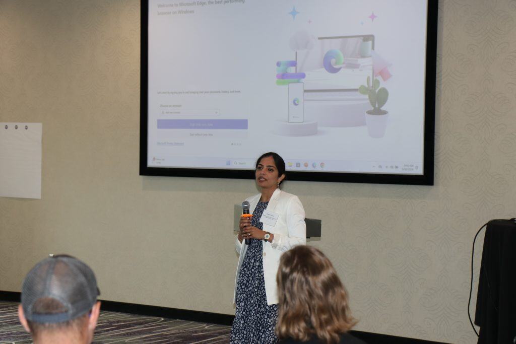 At the 2023 BioWRAP Annual Meeting, a woman in a white blazer speaks into a microphone before an audience, with a large screen displaying a Microsoft Edge welcome page behind her in the conference room. - BioWRAP