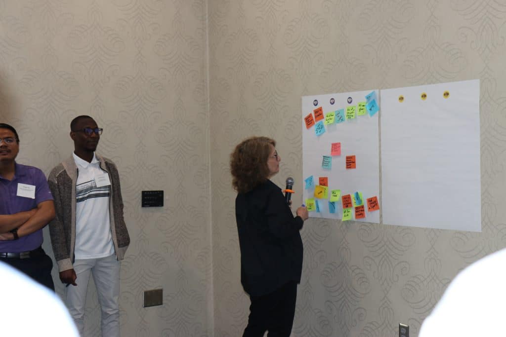A person holding a microphone stands near a wall with two large sheets of paper covered in colorful sticky notes, while two people stand nearby watching during the BioWRAP 2023 Annual Meeting workshop or group discussion. - BioWRAP