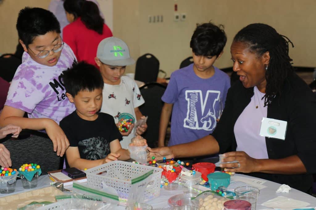 A woman and four children gather around a table covered with colorful craft supplies, making art projects together at the 2023 BioWRAP Annual Meeting. The group looks engaged and happy in a lively, indoor setting. - BioWRAP
