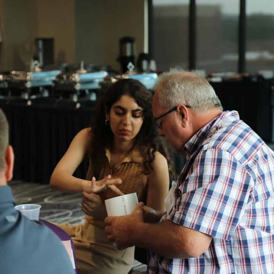 At the 2023 BioWRAP Annual Meeting, a woman and an older man sit at a table in conversation. The woman gestures with her hands as the man holds a piece of paper. Buffet trays and another attendee are visible in the background. - BioWRAP
