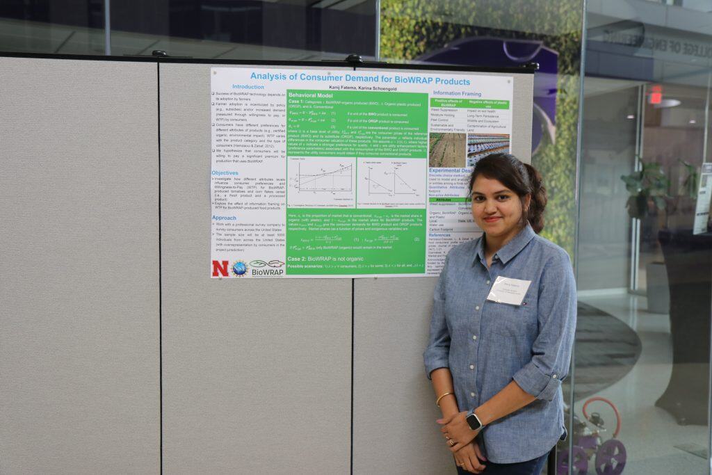 A woman stands and smiles next to a scientific poster titled “Analysis of Consumer Demand for BioWRAP Products” at the 2023 Annual Meeting, displayed on a grey board in an indoor setting. She is wearing a blue shirt and a name badge. - BioWRAP
