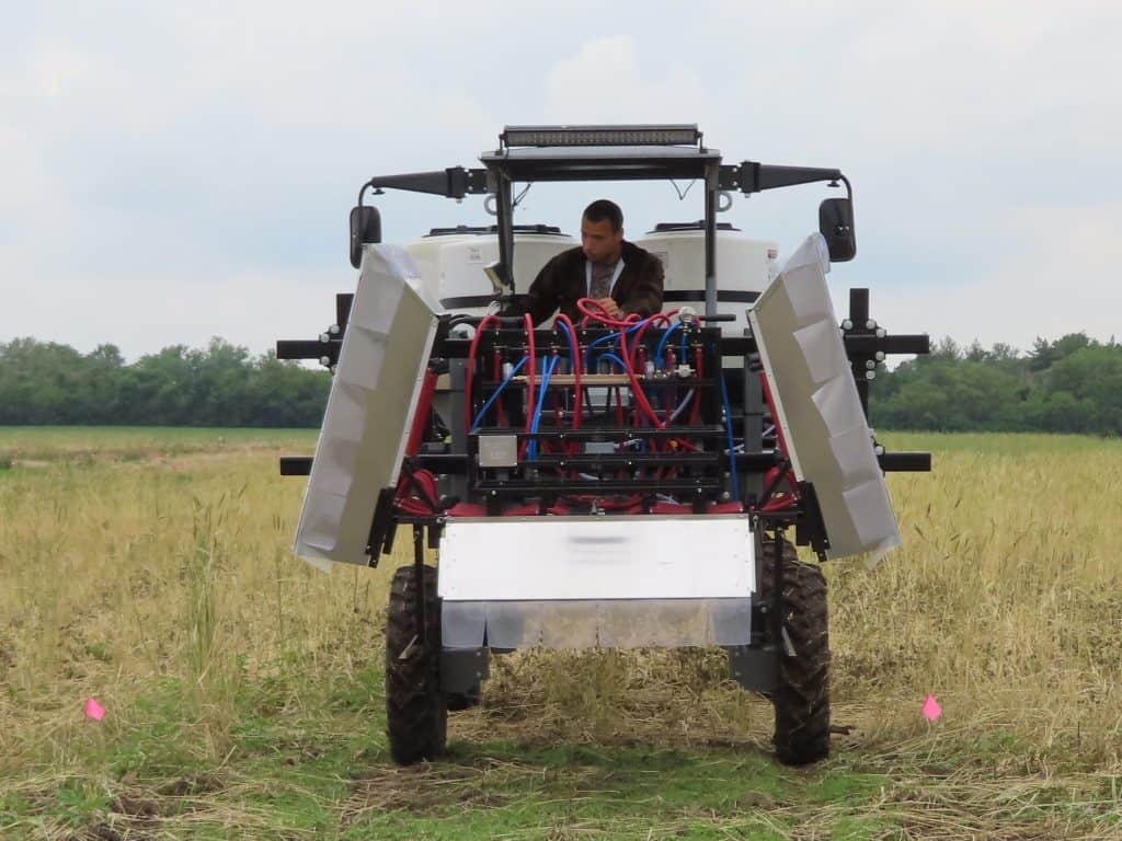 At the 2025 BioWRAP Annual Meeting, a person stands in a grassy field next to an off-road vehicle equipped with gear, while another person sits inside. Trees and a cloudy sky form the backdrop. - BioWRAP