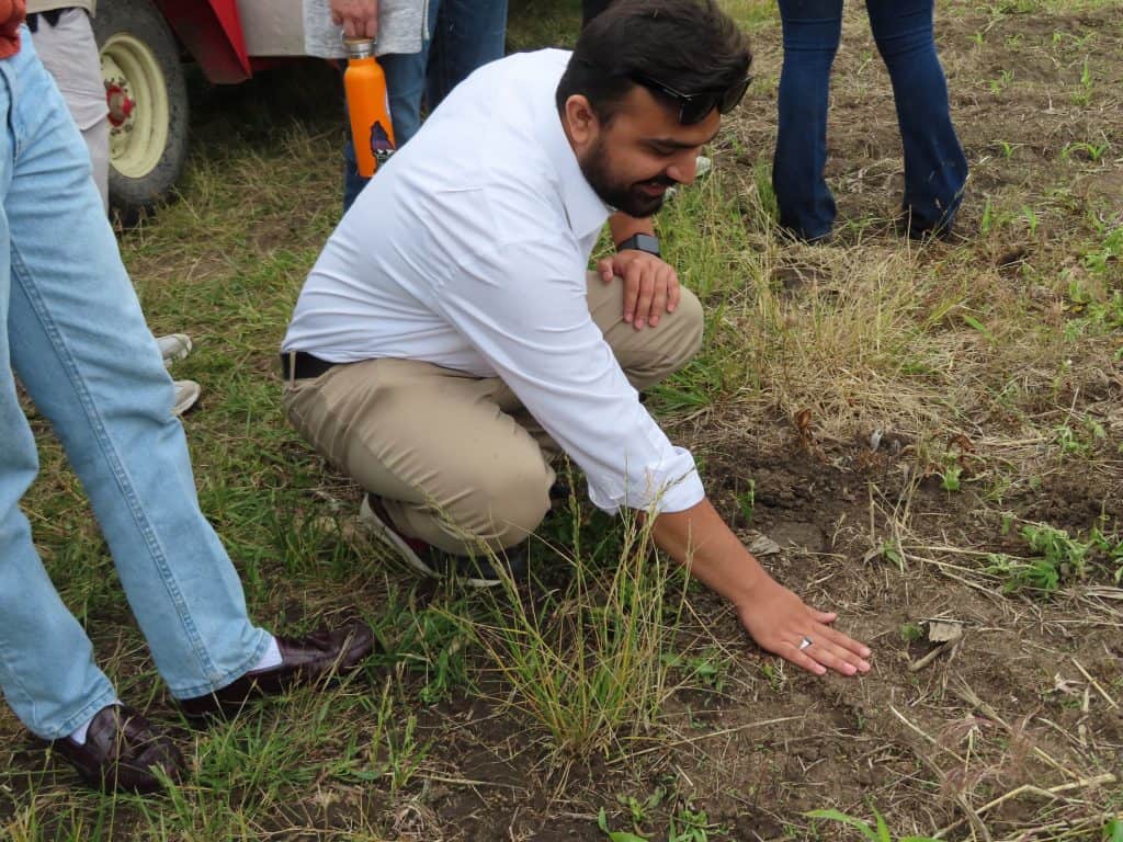 At the 2025 Annual Meeting, a man in a white shirt and khaki pants crouches on the ground, touching the soil with his hand in a field where other attendees are gathered among grass and plants. - BioWRAP