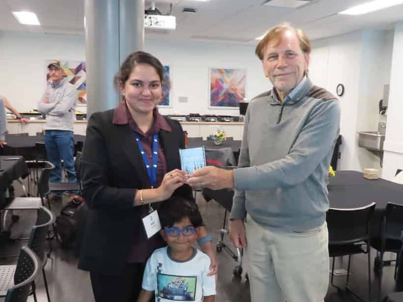 At the 2025 BioWRAP Annual Meeting, a woman receives an award from an older man in a conference room. A child stands smiling in front of them, while attendees sit at tables with black tablecloths in the background. - BioWRAP