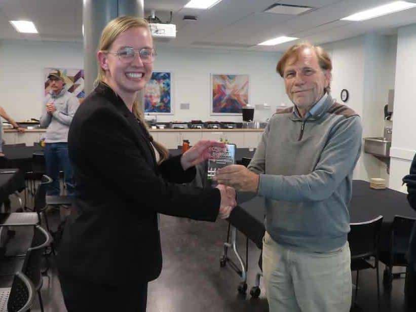 A smiling woman in a suit receives an award from an older man in a casual outfit at the BioWRAP 2025 Annual Meeting, while others stand and watch in a modern conference room. - BioWRAP
