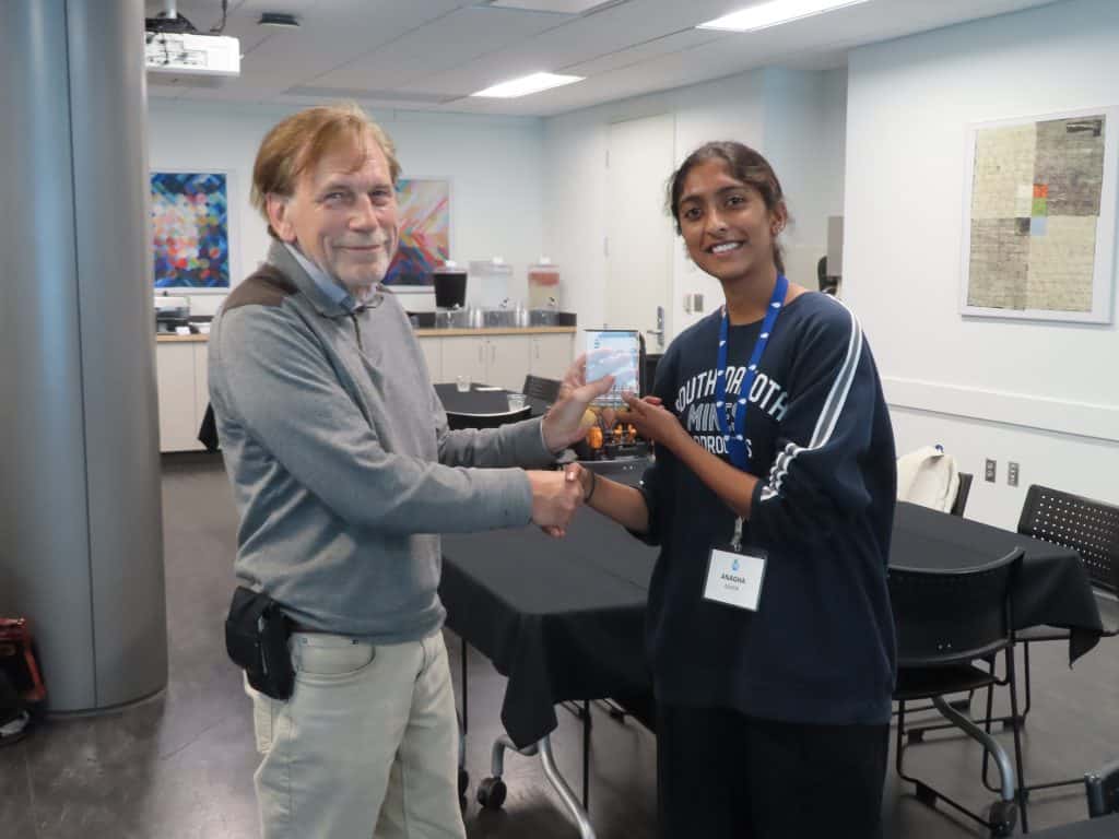 A man and a woman are shaking hands in a modern conference room at the 2025 BioWRAP Annual Meeting. The woman, wearing a name badge and lanyard, holds a small award as they both smile at the camera. Tables and chairs are in the background. - BioWRAP