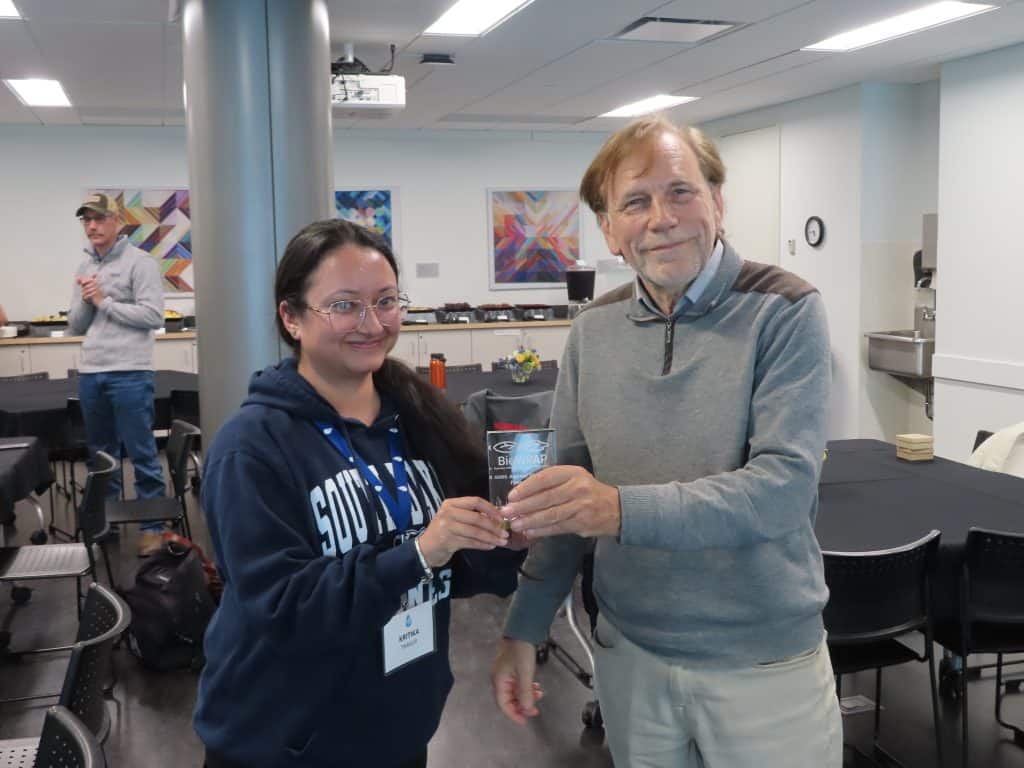 A woman and an older man smile as they hold a glass award together at the 2025 BioWRAP Annual Meeting in a brightly lit room with tables and abstract art on the walls. Other people and food trays are visible in the background. - BioWRAP