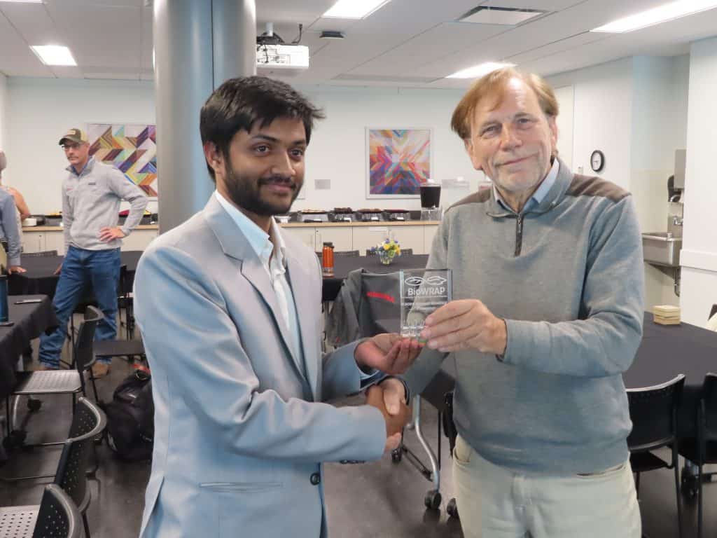 Two men are shaking hands while one presents a transparent BioWRAP award plaque to the other in a modern, well-lit conference room during the 2025 Annual Meeting. Other people and artwork are visible in the background. - BioWRAP