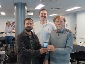 Three men stand together in a room, smiling at the camera while holding a BioWRAP award. Two men are in front and a taller man stands behind them. In the background, another person is near a table and colorful wall art at the 2025 Annual Meeting. - BioWRAP