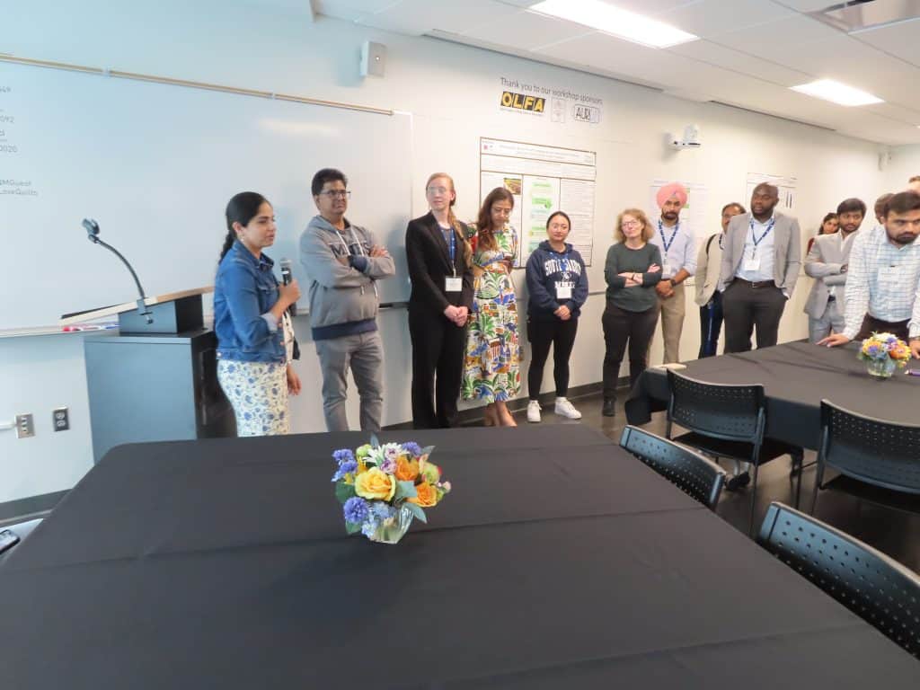 A diverse group of people stand in a classroom near a whiteboard and posters during the 2025 BioWRAP Annual Meeting. Tables with flower centerpieces are in the foreground, and most attendees wear nametags and business or casual attire. - BioWRAP