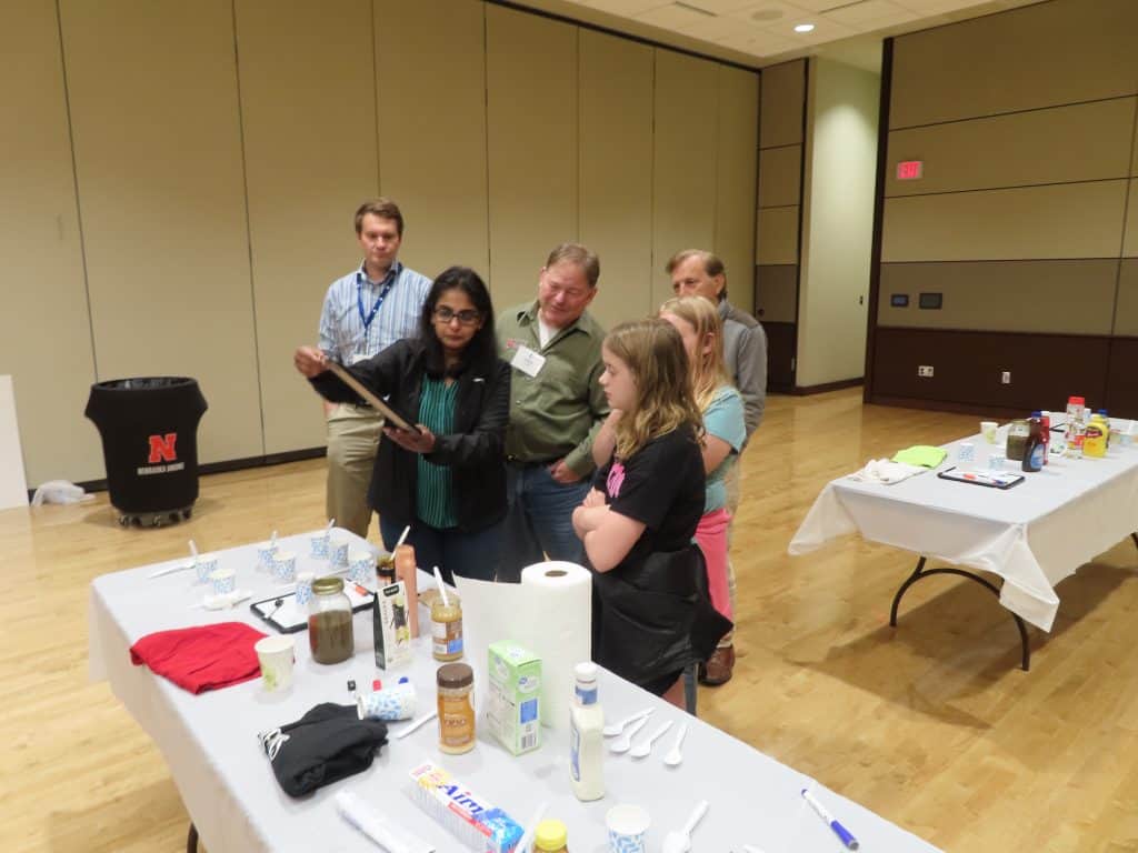 A group of people stand around a table covered with food items and supplies at the 2025 BioWRAP Annual Meeting, while a woman shows something on a tray to the others in a spacious, well-lit room with beige walls and a wooden floor. - BioWRAP