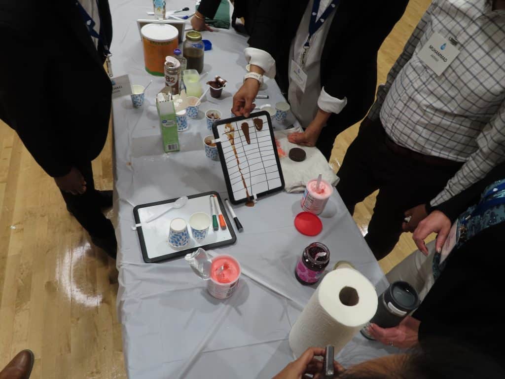 At the BioWRAP Annual Meeting 2025, people in business attire gather around a table filled with ice cream toppings, paper cups, napkins, and a whiteboard drizzled with syrup for a fun ice cream social or demonstration event. - BioWRAP