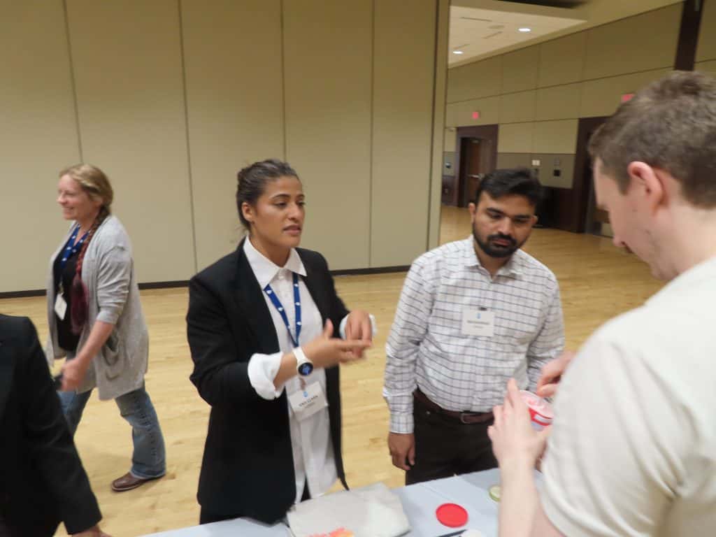 Four people are interacting at a table in a spacious conference room during the 2025 BioWRAP Annual Meeting. Two face the camera, engaged in conversation, while a third stands beside them and a fourth walks in the background. - BioWRAP