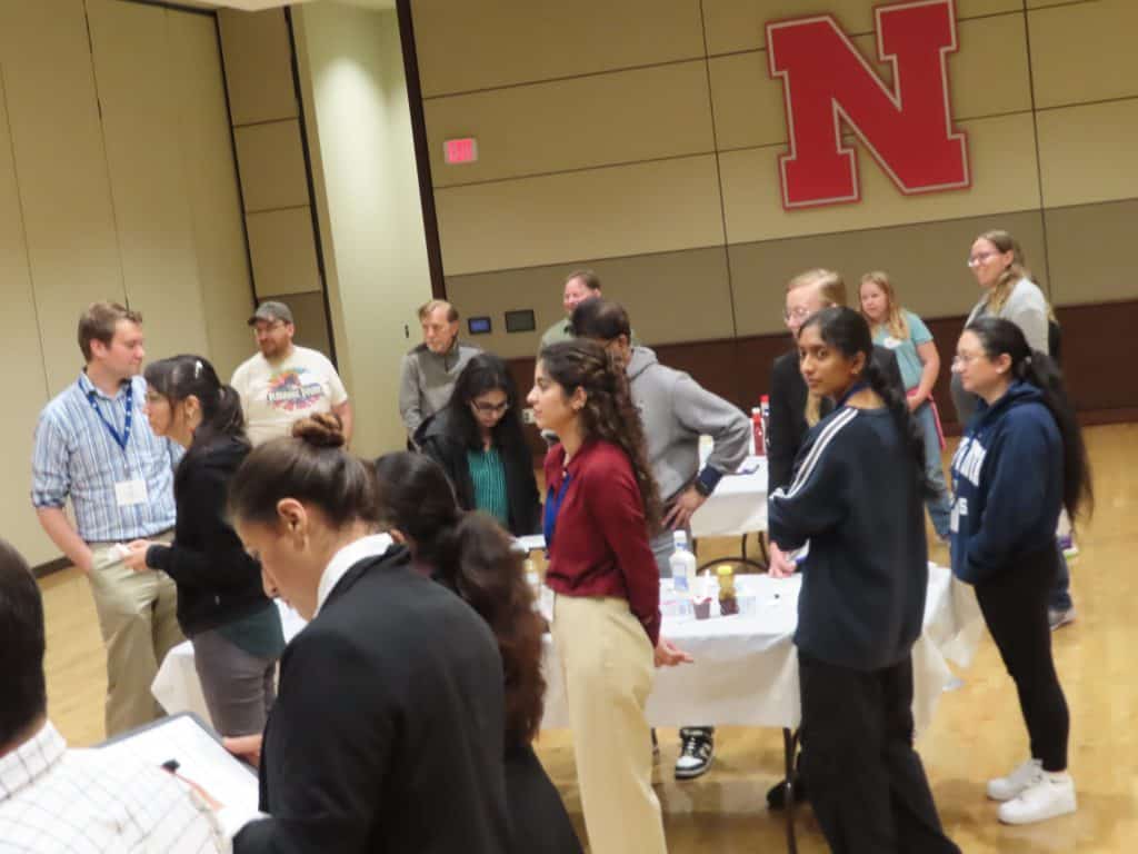 A group of people stand and converse around tables in a large room with a wooden floor. The red "N" logo on the wall signals the University of Nebraska, hosting the 2025 Annual Meeting for BioWRAP. - BioWRAP