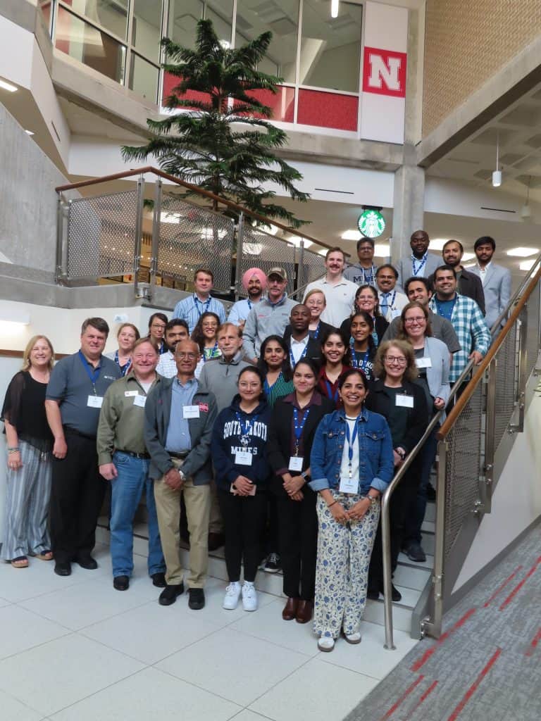 A large, diverse group of adults stands smiling on a staircase and landing in a modern building with a tree, metal railings, and a University of Nebraska "N" sign, gathered for the 2025 BioWRAP Annual Meeting. - BioWRAP