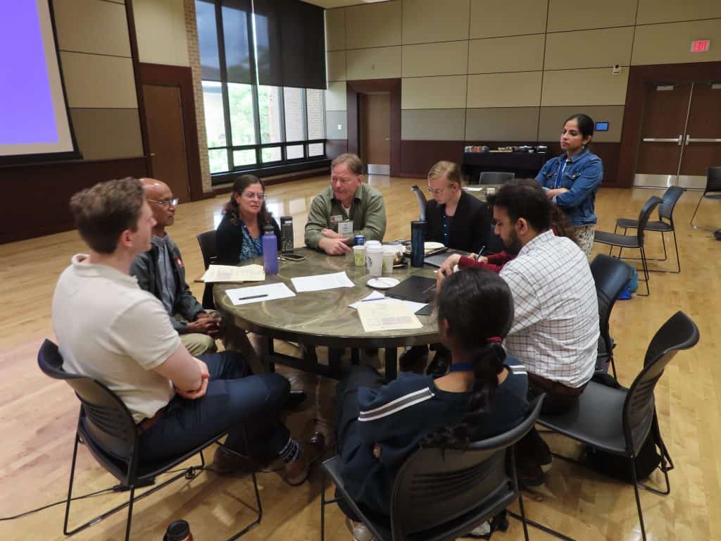 A group of eight people sit and stand around a round table in a large, well-lit room, engaged in discussion at the BioWRAP 2025 Annual Meeting, with papers, notebooks, and drinks on the table. - BioWRAP