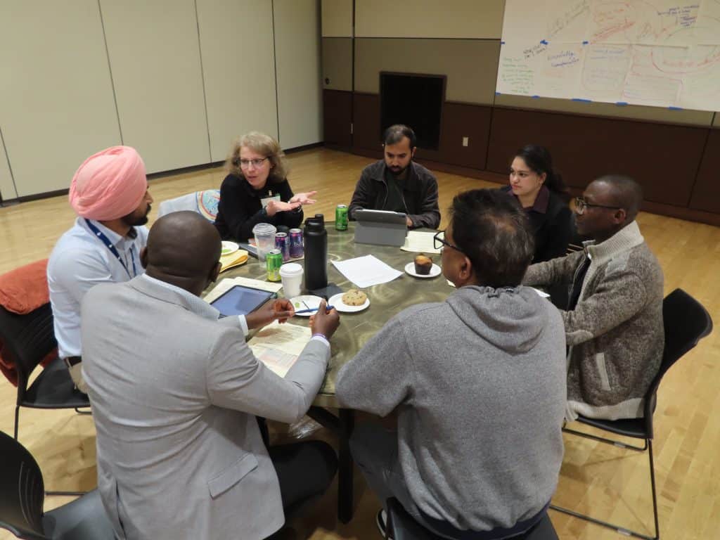Six adults sit around a round table in discussion, some using tablets and laptops. Papers, notebooks, drinks, and snacks are on the table. A whiteboard with notes is in the background at the BioWRAP 2025 Annual Meeting. - BioWRAP