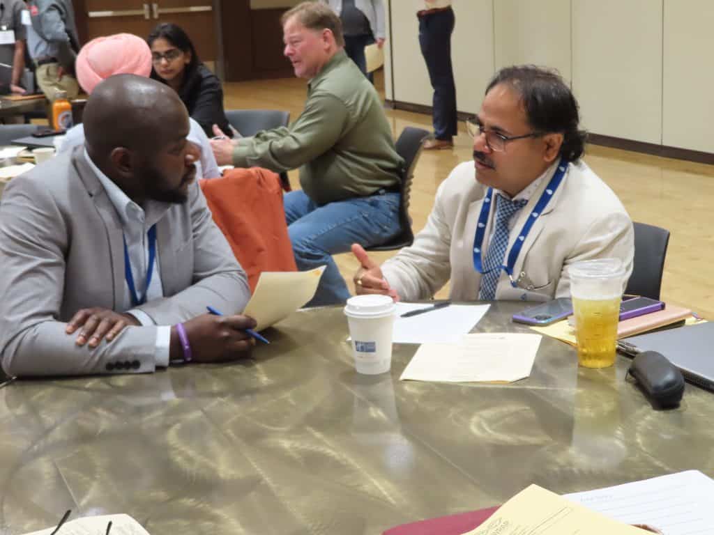 Two men in business attire sit at a table during the 2025 Annual Meeting, discussing BioWRAP documents. One gestures while the other listens. Several drinks, folders, and papers are on the table, with other attendees talking in the background. - BioWRAP