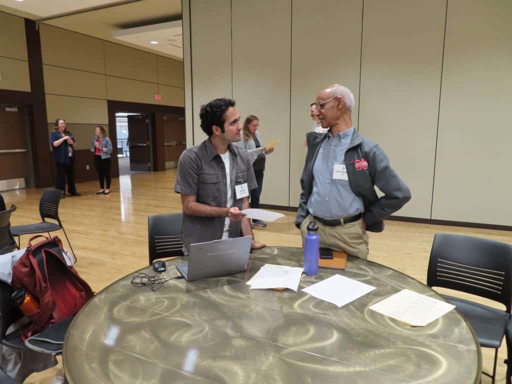 Two people stand and talk at a round table with papers, a water bottle, and a laptop during the 2025 BioWRAP Annual Meeting. Others are gathered in the background, suggesting an engaging workshop or collaborative session. - BioWRAP