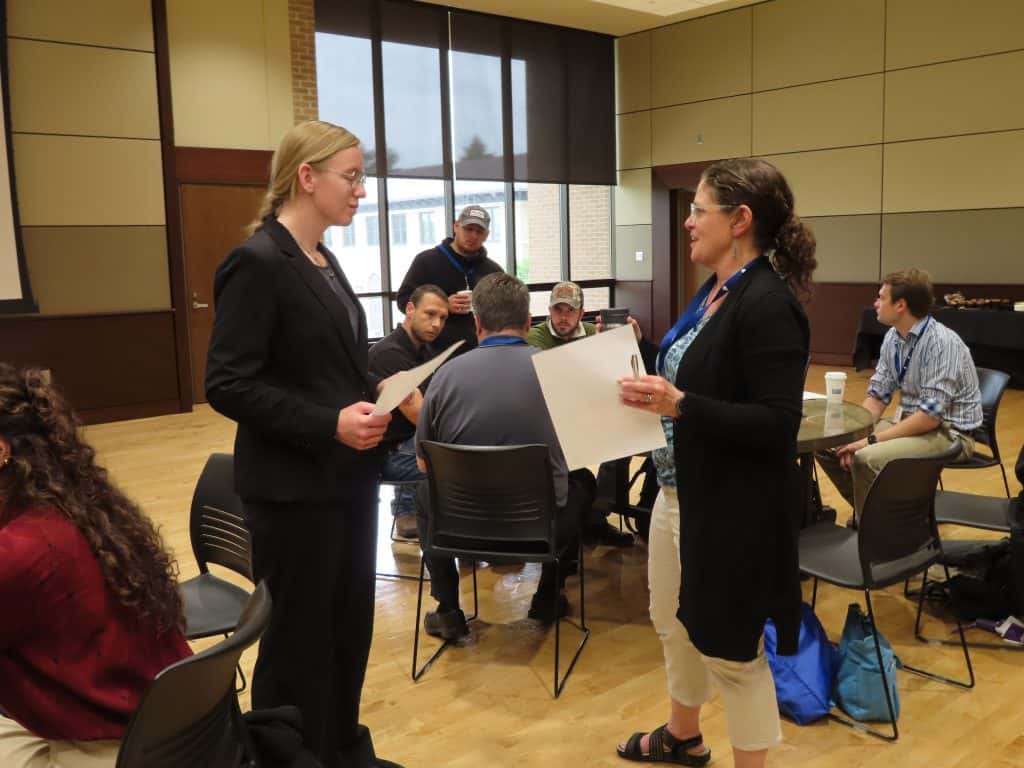 Two women stand and talk while holding papers in a meeting room during the 2025 BioWRAP Annual Meeting, as several people sit and converse at tables in the background. The setting is casual and collaborative. - BioWRAP