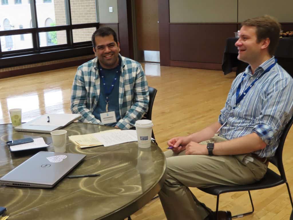 Two men sit at a round table with papers, a laptop, and coffee cups, smiling and talking about the 2025 BioWRAP Annual Meeting in a bright room with wooden floors and large windows. - BioWRAP