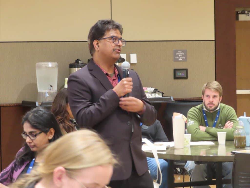 At the Annual Meeting 2025, a man in a suit stands and speaks into a microphone at an indoor event, while others sit and listen around him at tables. - BioWRAP