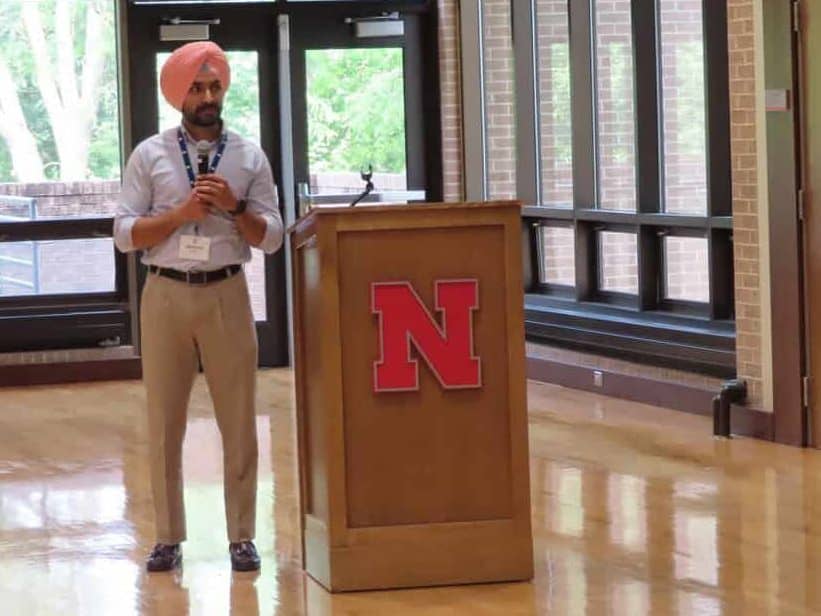 A man wearing a pink turban speaks into a microphone behind a wooden podium with a red "N" logo in a bright room at the University of Nebraska during the BioWRAP Annual Meeting 2025. - BioWRAP
