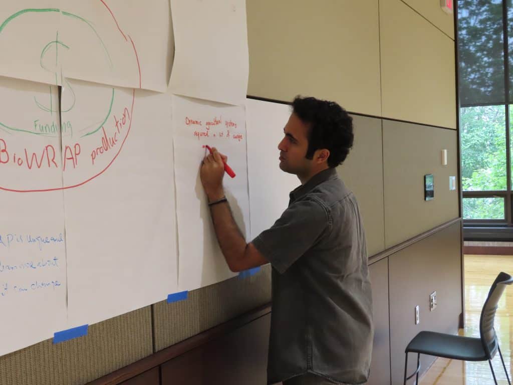 A man stands next to large sheets of paper taped to a wall, writing on one with a red marker during what appears to be a group brainstorming session at the BioWRAP Annual Meeting. - BioWRAP