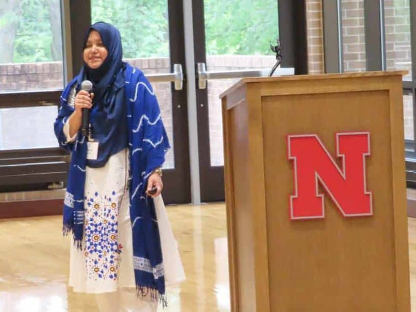 A woman in a blue hijab and white dress speaks into a microphone at the 2025 BioWRAP Annual Meeting, standing next to a podium with a large red "N" logo in a bright room with large windows and polished wooden floors. - BioWRAP