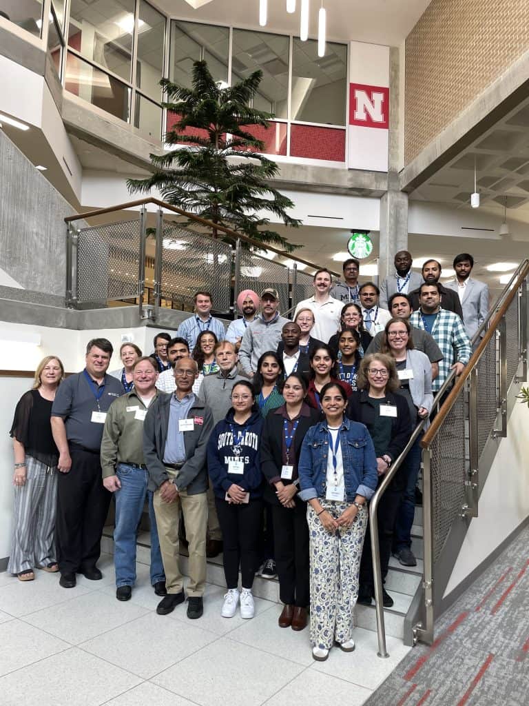 A group of about 30 people poses for a photo on a staircase indoors at the Annual Meeting, with a University of Nebraska–Lincoln banner and a Starbucks sign in the background. The diverse group represents BioWRAP’s 2025 gathering. - BioWRAP
