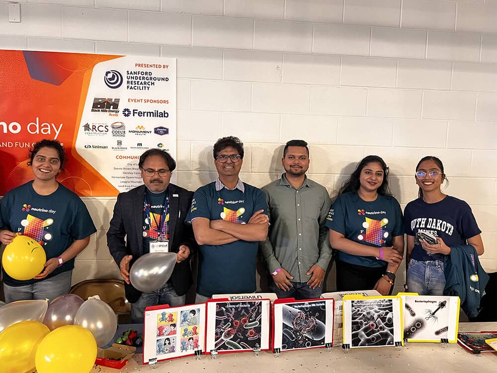 Six people stand smiling behind a table displaying science-themed images and balloons. Four wear matching dark t-shirts, one wears a jacket, and one wears a gray shirt. A colorful South Dakota Mines event banner hangs behind them on a white wall. - BioWRAP