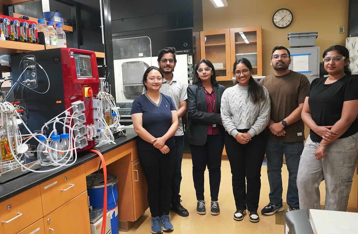 Six people stand together in a laboratory surrounded by cutting-edge equipment, shelves, and workstations. They are posing for a group photo and appear to be part of a research team making significant research advancements. - BioWRAP